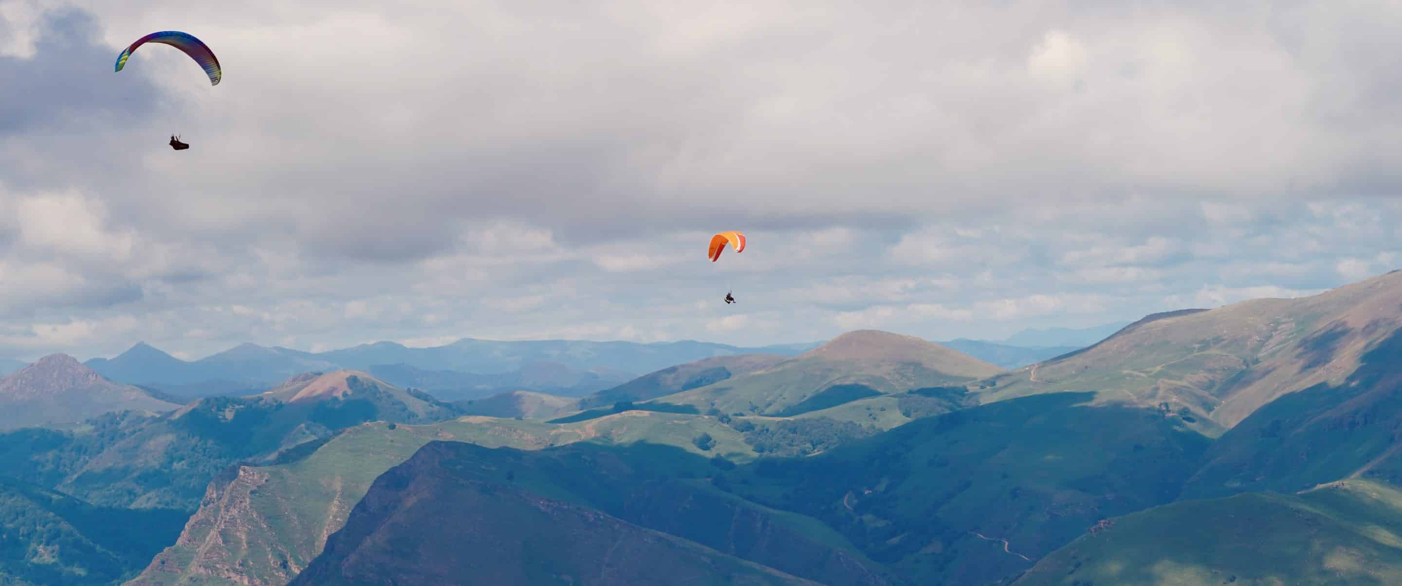 Deux parapentes survolant les paysages verdoyants et vallonnés du Mont Baigura, dans les Pyrénées-Atlantiques, sous un ciel nuageux.