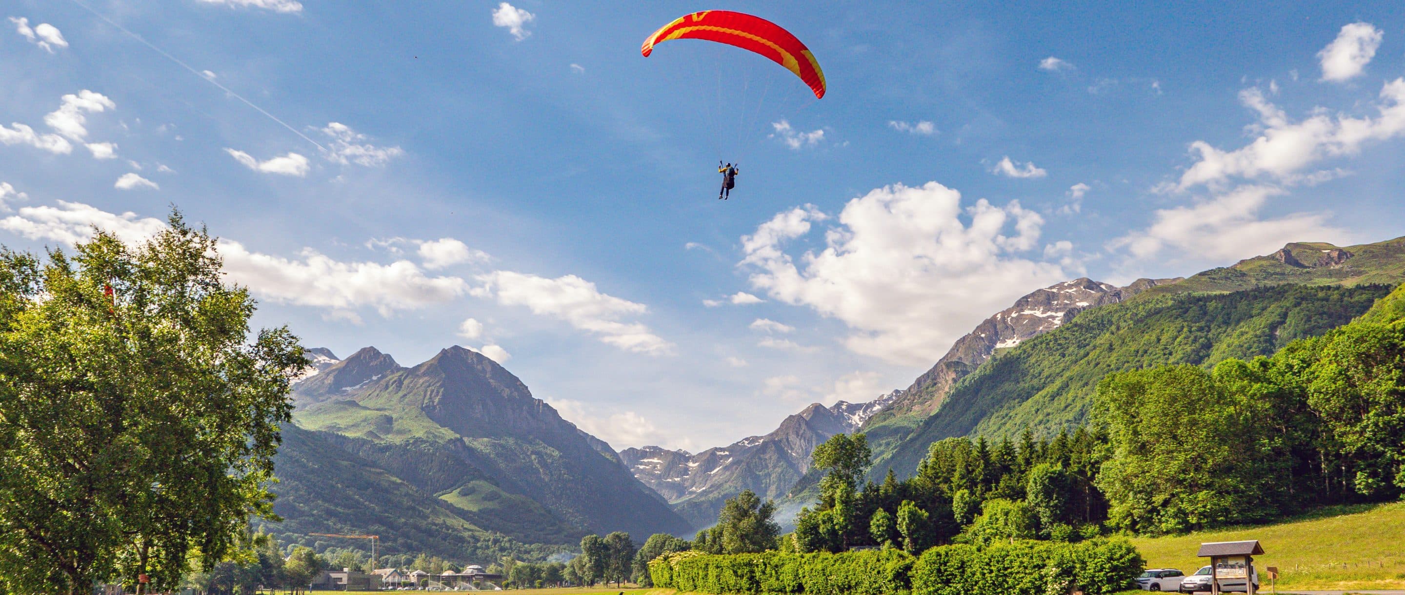 Un parapente rouge en vol au-dessus de Val Louron, dans les Hautes-Pyrénées, avec des montagnes verdoyantes en arrière-plan sous un ciel dégagé