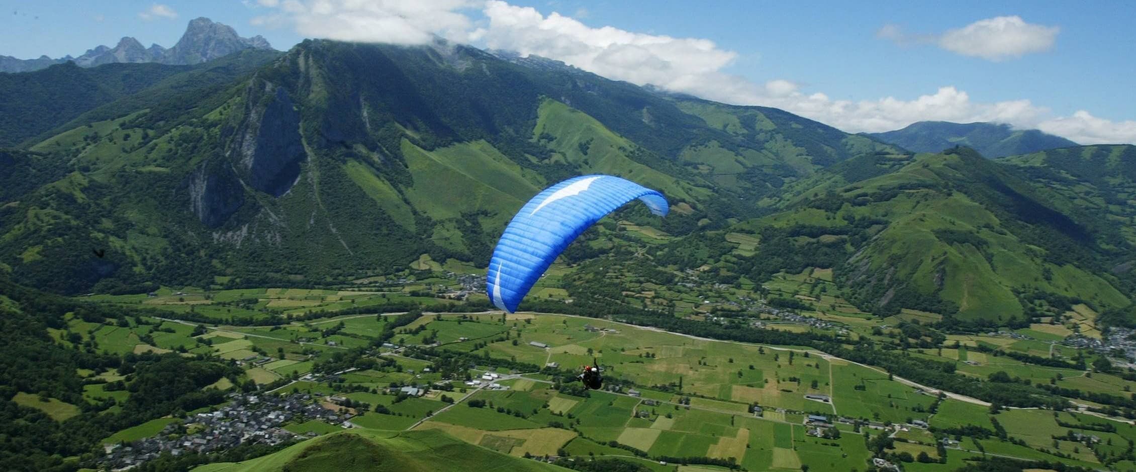 Un parapente bleu en vol au-dessus des paysages verdoyants de la vallée d'Accous, dans les Pyrénées-Atlantiques, avec des montagnes en arrière-plan.